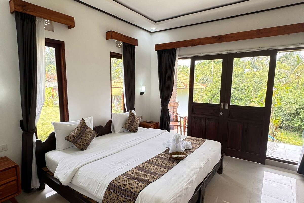 A spacious bedroom with a large bed, wooden headboard, and a view of lush greenery through the sliding glass doors.