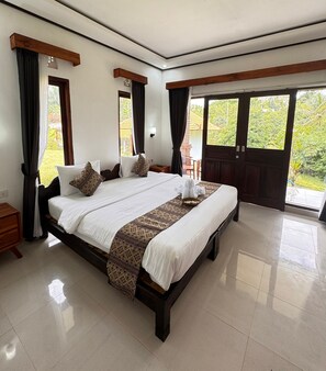 A spacious bedroom with a large bed, wooden headboard, and a view of lush greenery through the sliding glass doors. - Indrayoga Retreat Bali (Selemadeg)