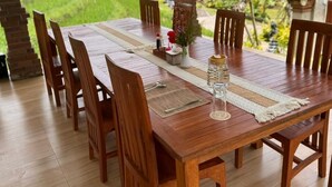 Une table à manger en bois, accompagnée de chaises, un vase rempli de fleurs et un verre d’eau, le tout placé devant un paysage verdoyant et luxuriant, parsemé de palmiers.