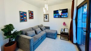 Een woonkamer met een grijze hoekbank, een grote flatscreen televisie aan de muur, een potplant en een schuifdeur van glas.