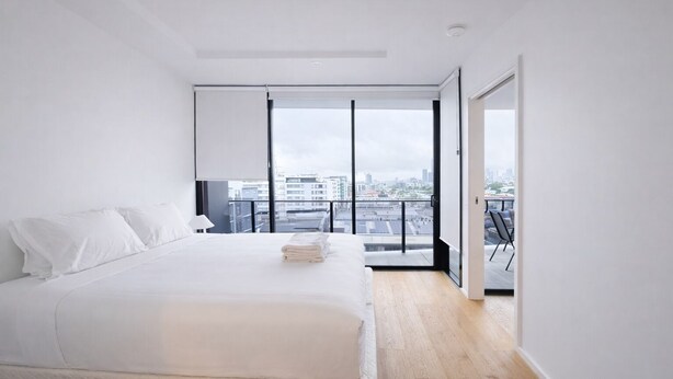 A modern bedroom with a large bed, a view of the city through floor-to-ceiling windows, and a balcony with a cityscape view. - 2BR Aprt West End Hideaway | City Views and Carpark (West End)