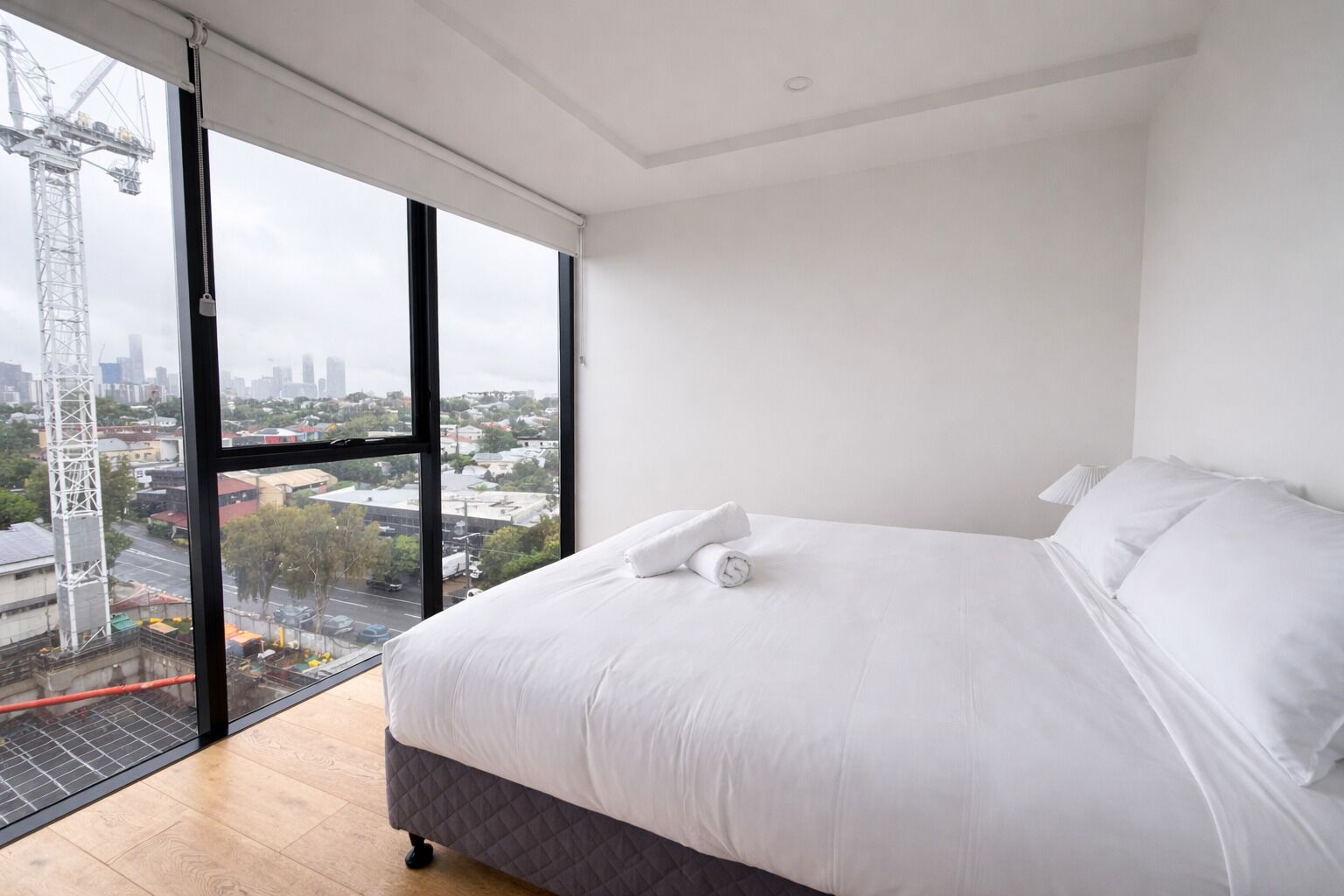 A modern bedroom with a large bed, white linens, and a view of a cityscape through a large window.