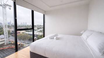 A modern bedroom with a large bed, white linens, and a view of a cityscape through a large window.