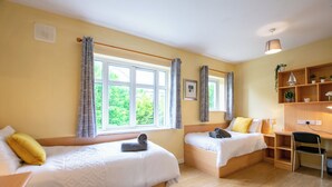 Ein Zimmer mit zwei Betten, einem Schreibtisch und einem Stuhl. Fenster mit Vorhängen, ein Regal mit Pflanzen und einer Wandleuchte.
