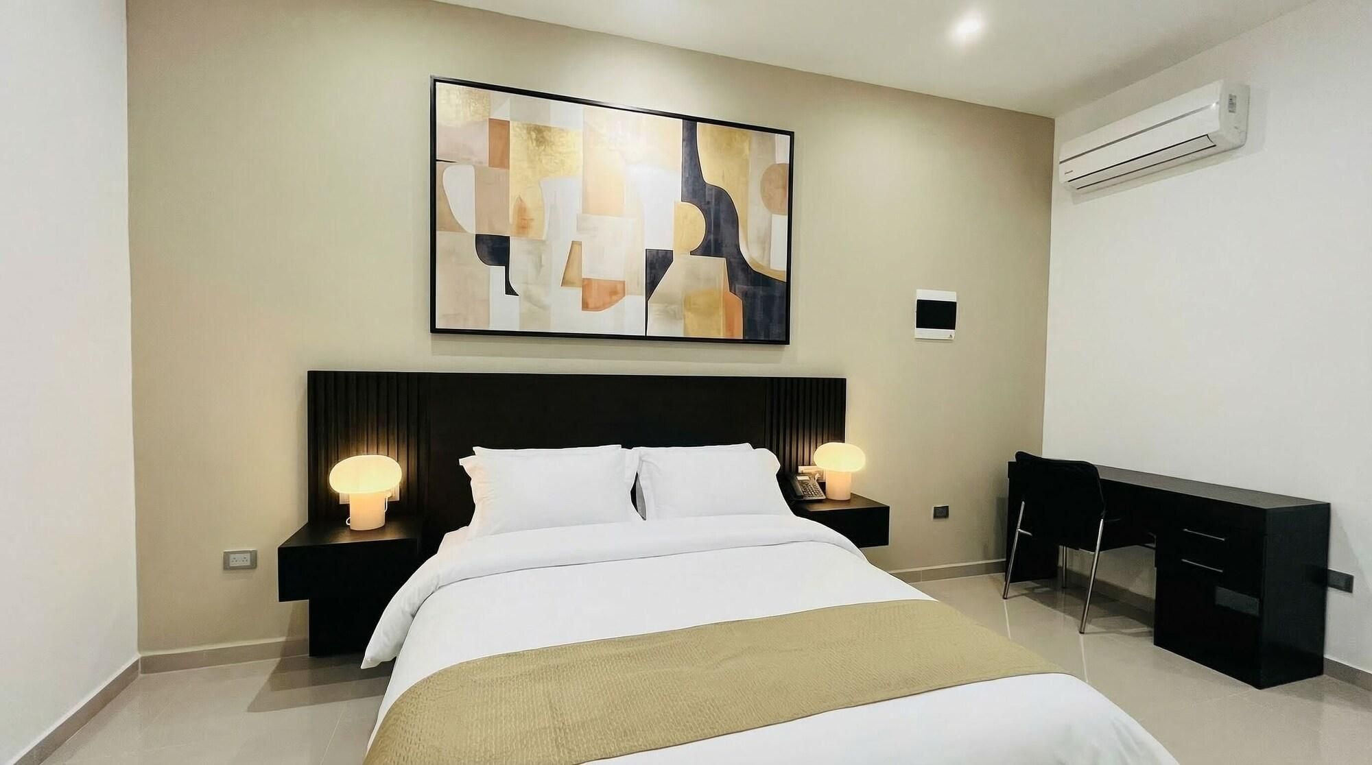 A modern hotel room with a bed, bedside lamps, a desk, and an abstract painting on the wall.