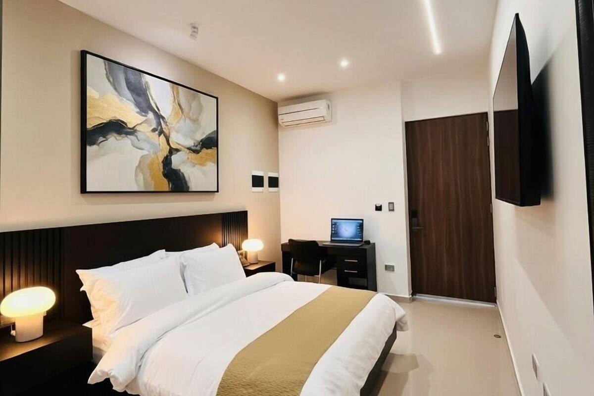 Ein modernes Hotelzimmer mit einem großen Bett, einem Schreibtisch mit einem Laptop, einem Flachbildfernseher und einer Klimaanlage.
