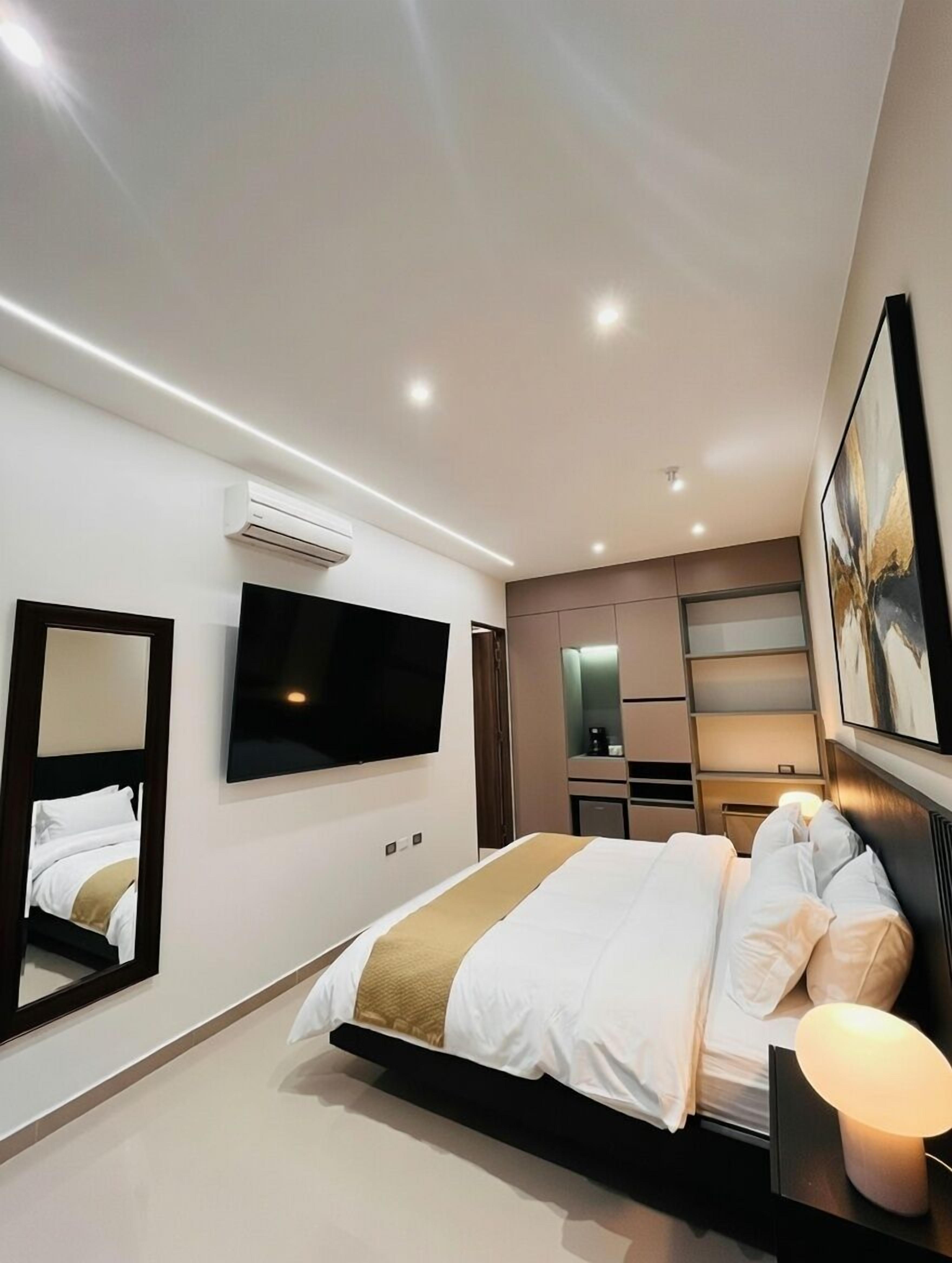 Une chambre d’hôtel moderne équipée d’un grand lit, d’un téléviseur à écran plat, d’un miroir, d’une table de chevet avec une lampe et d’un climatiseur fixé au mur.