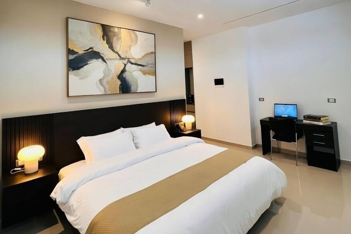 Ein modernes Hotelzimmer mit einem großen Bett, einem Schreibtisch mit Computer und einem an der Wand hängenden abstrakten Gemälde.
