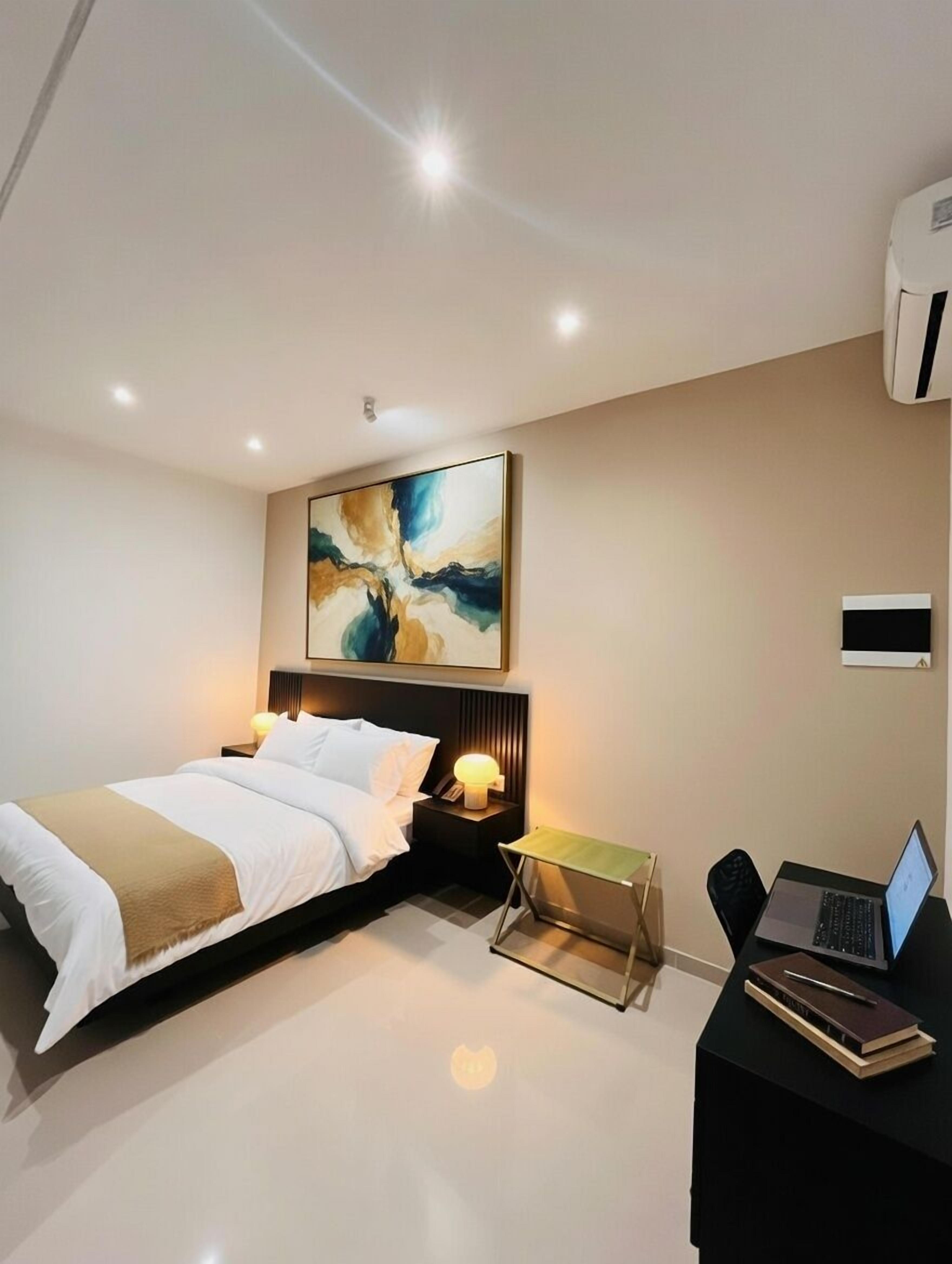 Une chambre d’hôtel moderne, équipée d’un lit, de tables de chevet, d’un bureau avec un ordinateur portable et d’un grand tableau abstrait accroché au mur.