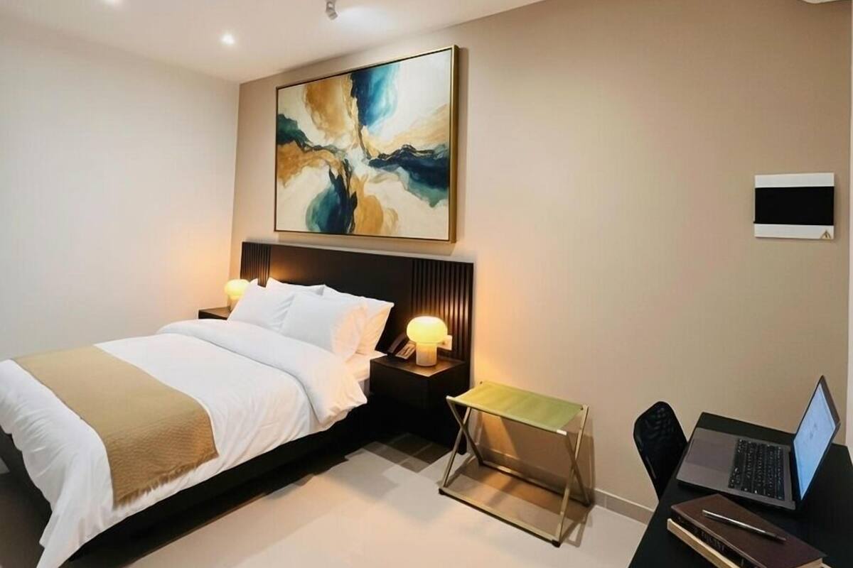 Ein modernes Hotelzimmer mit einem Bett, Nachttischen, einem Schreibtisch mit einem Laptop und einem großen, abstrakten Gemälde an der Wand.