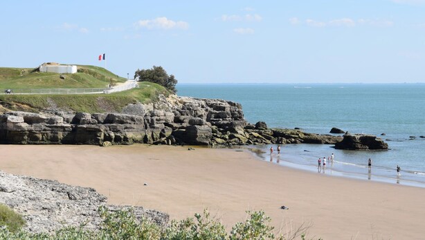 Ein Küstenstrand mit felsigen Klippen, ein Weg zu einem Gebäude und freie Sicht auf das Meer.
