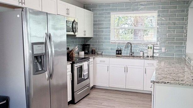 Una cocina moderna con armarios blancos, electrodomésticos de acero inoxidable, horno empotrado y ventana con vista a árboles.