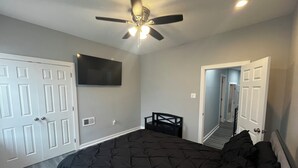 Ein Schlafzimmer mit einem Bett, einem Deckenventilator, einem an der Wand befestigten Fernseher und einer Tür zu einem weiteren Raum.