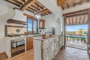 A kitchen with a stone island, wooden beams, a view to the outside, and a dining area with a table set for a meal. - Casale Le Sorgenti - Farmhouse with Pool for 8, Pitigliano, Italy (Pitigliano)