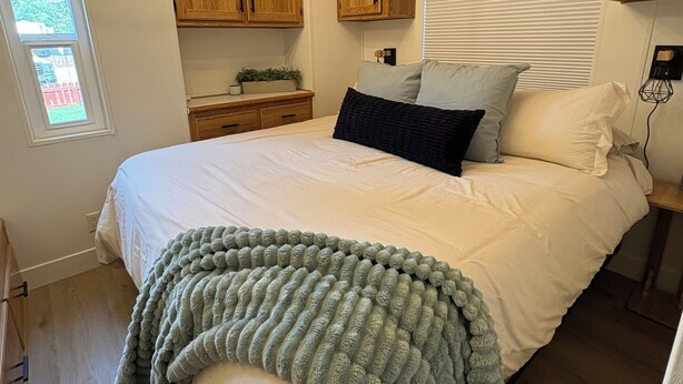 A bedroom with a large bed, wooden cabinets, a chandelier, and a window with blinds. - Cozy Cascade Cottage (Cascade)