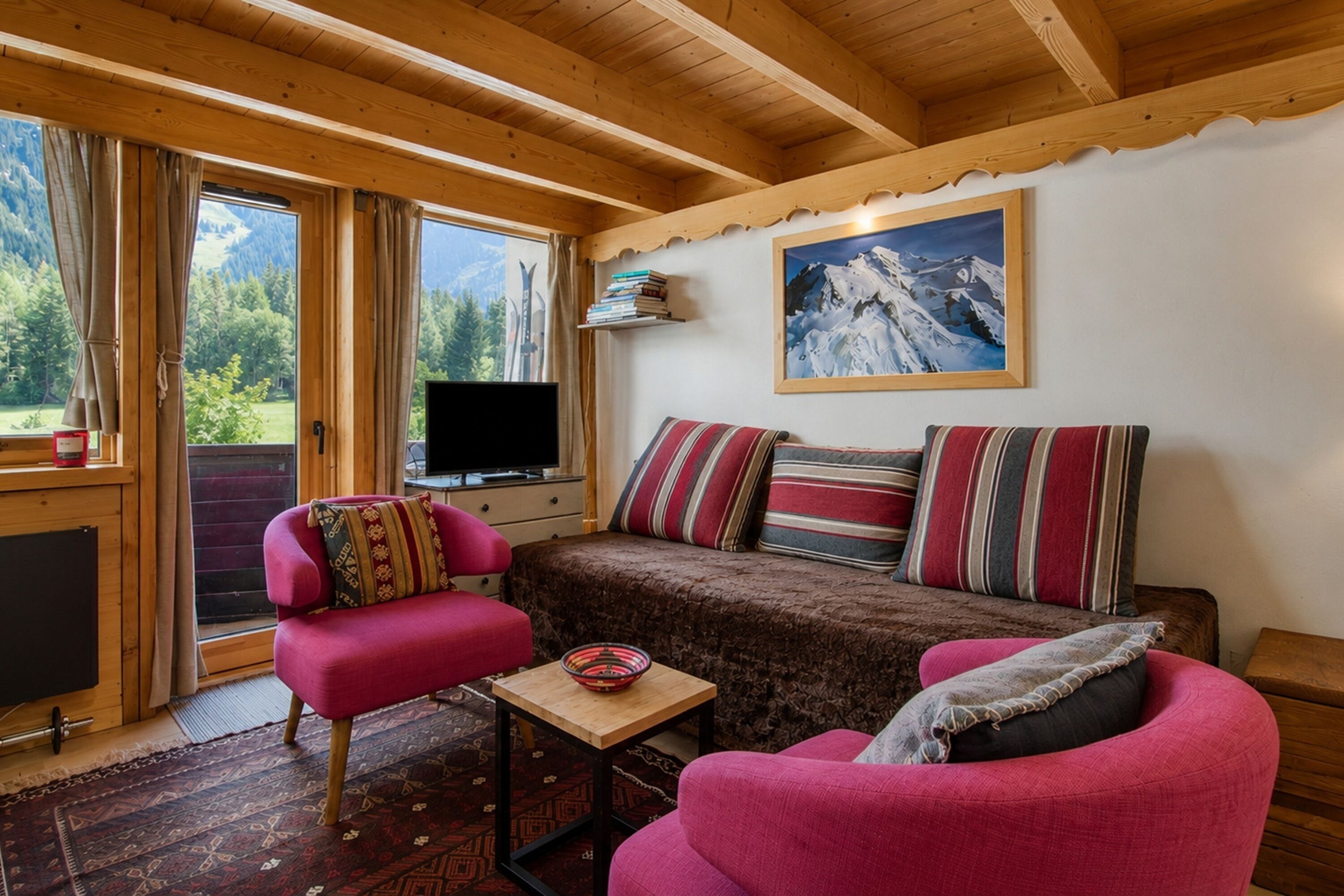 A cozy cabin interior with a wooden ceiling, a sofa with striped cushions, a pink armchair, a television, and a mountain view through the window.