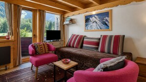 L’intérieur d’un chalet chaleureux, avec un plafond en bois, un canapé orné de coussins à rayures, un fauteuil rose, un téléviseur et une vue sur les montagnes par la fenêtre.