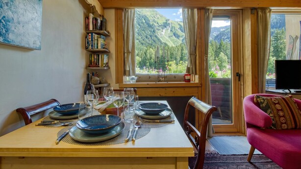 Une table à manger dressée avec des assiettes, des verres et des couverts, une bibliothèque, un canapé et une vue sur les montagnes par la fenêtre.