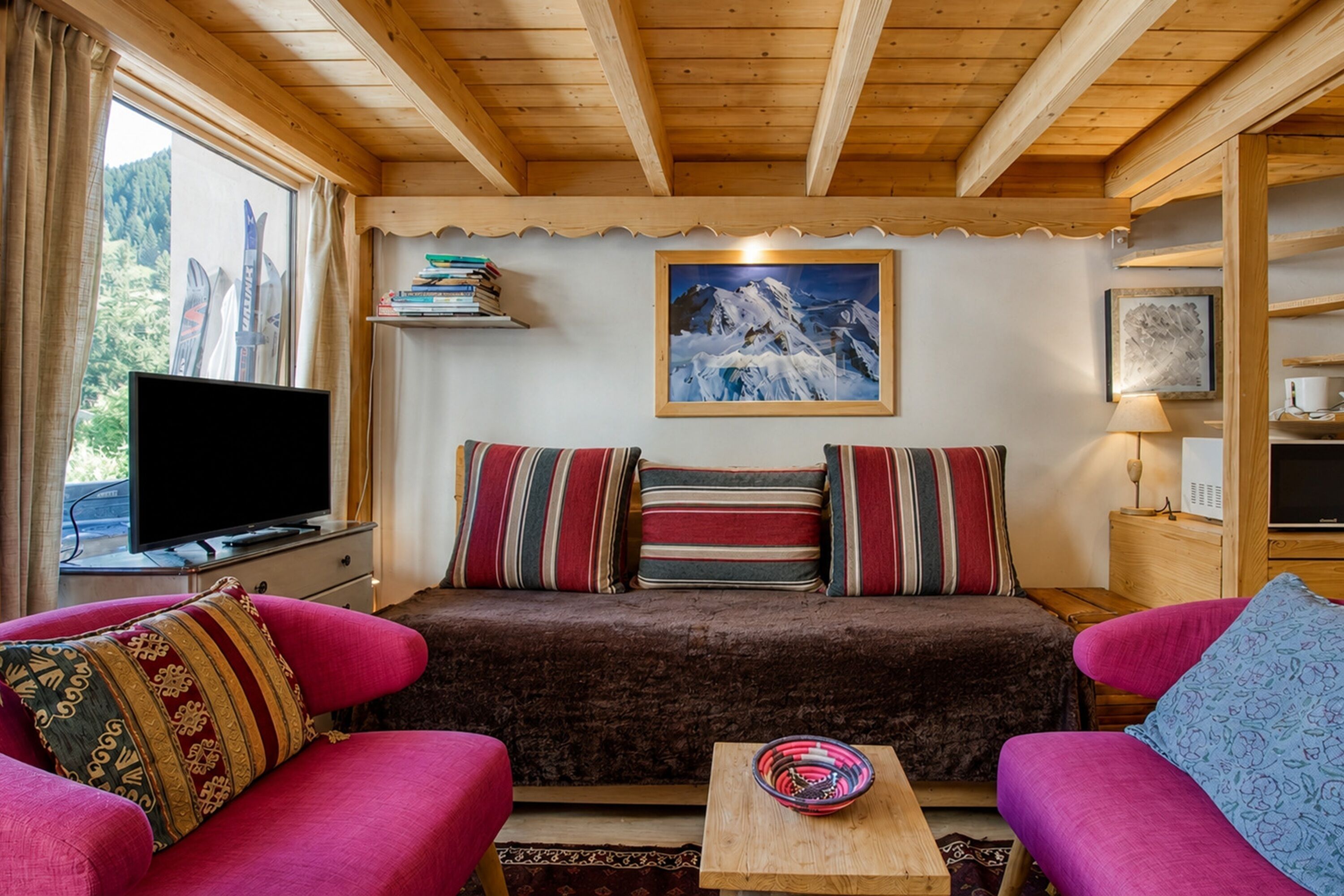 A cozy room with a wooden ceiling, a sofa with striped cushions, a television, and a framed picture of a mountain.