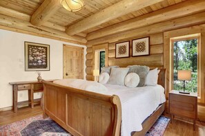 Un dormitorio de madera con una cama, mesitas de noche, una mesita auxiliar, una puerta y cuadros en la pared.