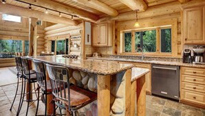 A log cabin kitchen with a stone island, bar stools, and a view of the forest.