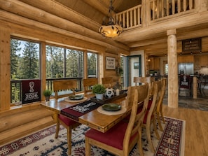 Un coin repas en bois, avec une table dressée pour quatre personnes, un lustre et une vue sur la forêt grâce à de grandes fenêtres.