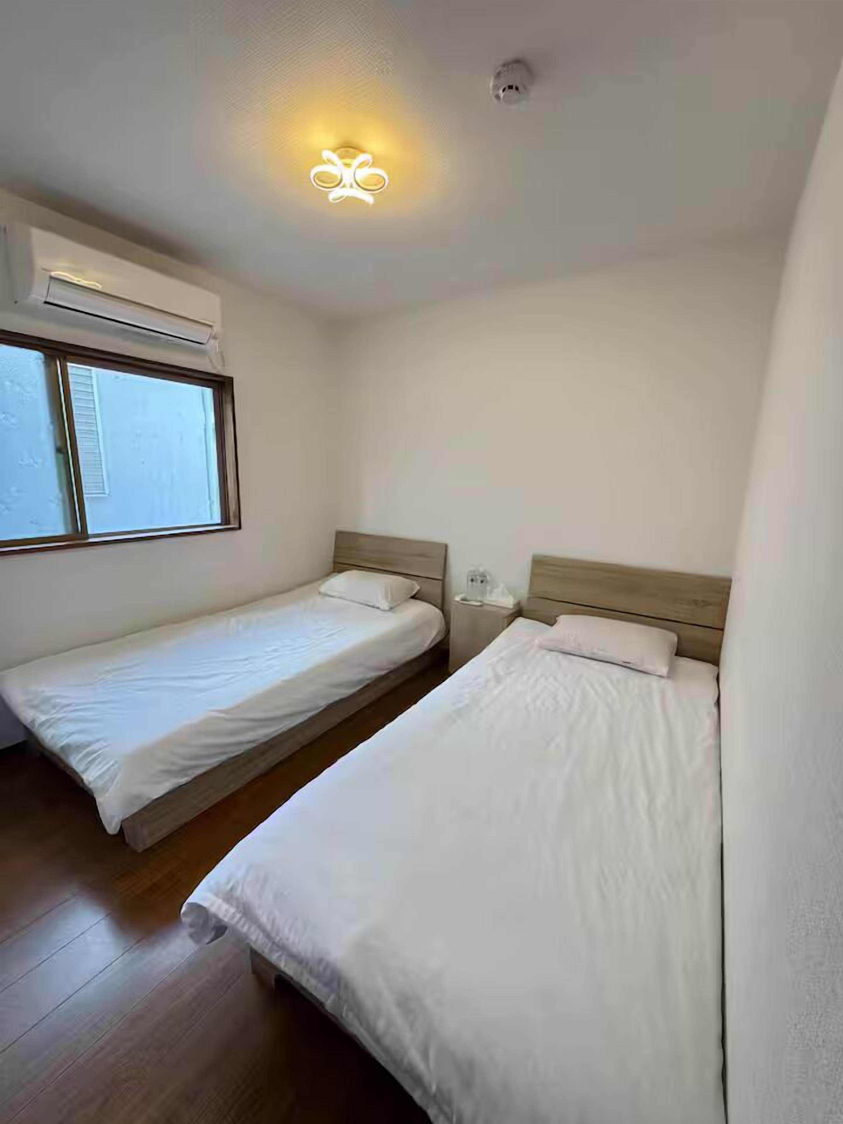 A small, clean room with two single beds, a window, and a ceiling light fixture.