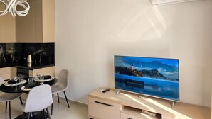 Ein modernes Wohnzimmer mit Flachbildfernseher, einem Essbereich mit Tisch und Stühlen sowie einer Küche mit schwarzen Geräten.