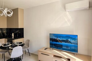 Ein modernes Wohnzimmer mit Flachbildfernseher, einem Essbereich mit Tisch und Stühlen sowie einer Küche mit schwarzen Geräten.