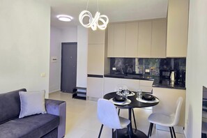 Ein modernes Wohnzimmer mit einer Couch, einem Esstisch für zwei Personen und einer Küchenzeile mit Schränken und Geräten.