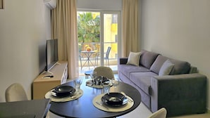 Ein modernes Wohnzimmer mit Esstisch für zwei Personen, einer Couch, einem Fernseher und einem Balkon mit Blick auf eine Palme.