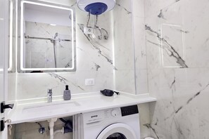 Une salle de bain avec un lavabo en marbre blanc, un miroir, une douchette à main et un lave-linge encastré.