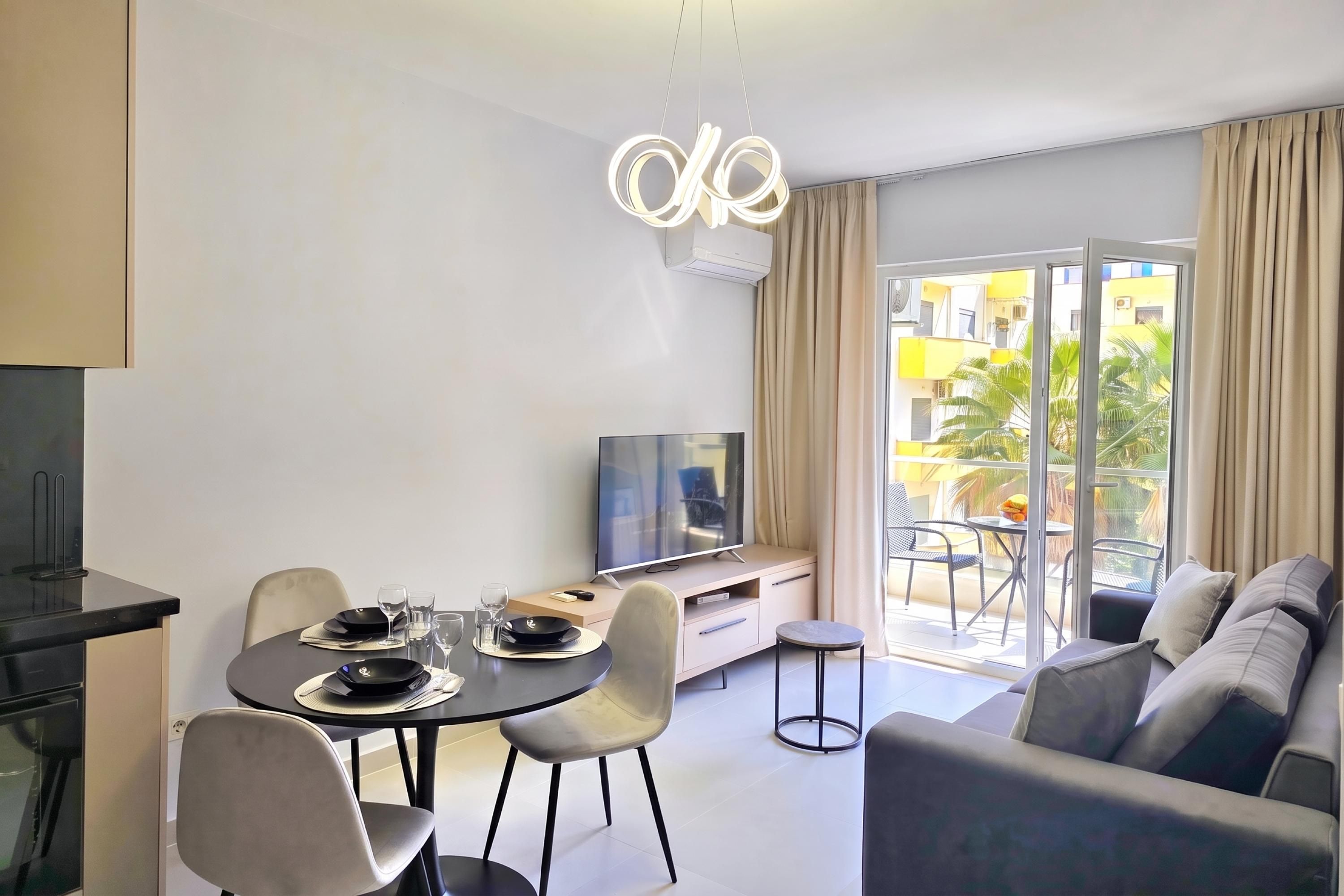 A modern living room with a dining area, a flat-screen TV, and a balcony with a view of the city.