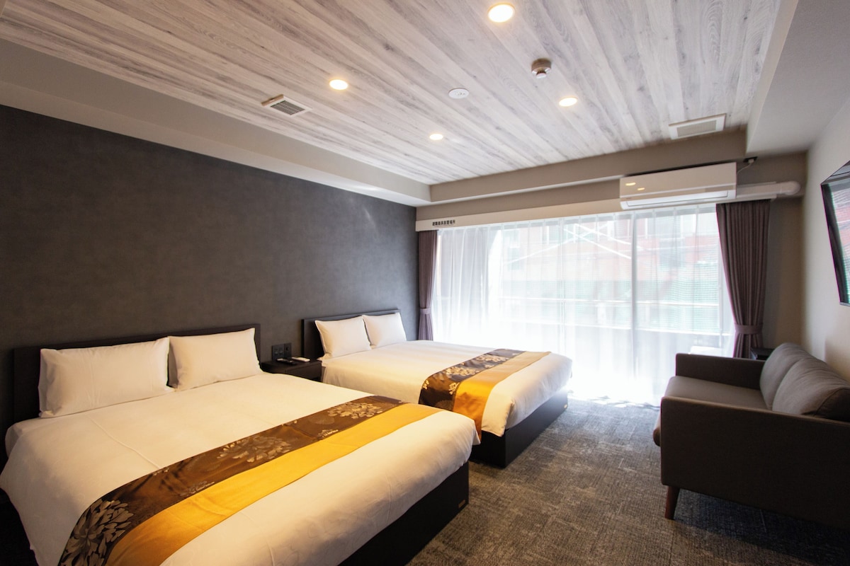 A hotel room with two beds, a sofa, a large window with curtains, and a ceiling with recessed lighting.