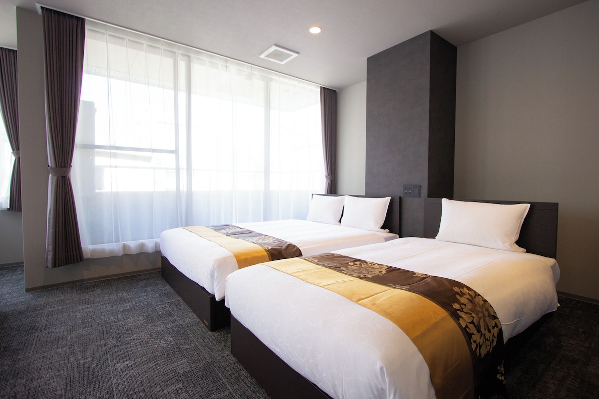 A hotel room with two beds, a large window with curtains, a carpeted floor, and a modern design.