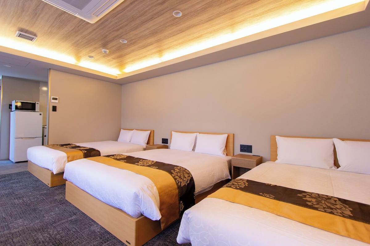 A hotel room with three single beds, each with a wooden headboard and a brown decorative bedspread. There is a small bedside table with a lamp on each bed. The room has a wooden ceiling with recessed lighting and a visible air conditioning unit on the left.