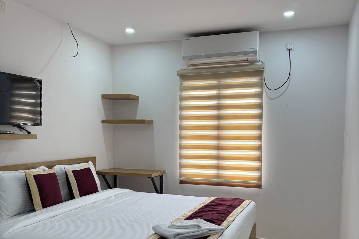 A neatly arranged bedroom with a bed, a TV, a shelf, and a window with blinds.