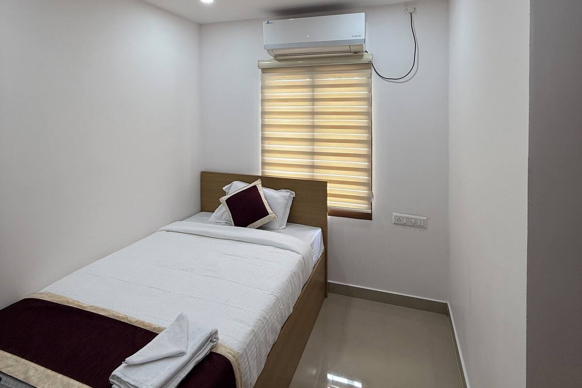 A hotel room with a bed, a window with blinds, an air conditioning unit, and a small bedside table.