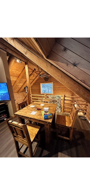 A wooden dining table with chairs, a television, and a window in a rustic room with wooden beams. - The Waterwheel Cabin & Guide Room/Family Retreat (Chiloquin)