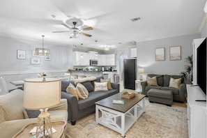A living room with a sofa, coffee table, and a TV. There is a kitchen in the background with cabinets and appliances. - Navarre Sound Place (Navarre)