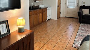 Sala de estar con televisor, armarios de madera, una zona de cocina con microondas y una puerta con persianas.