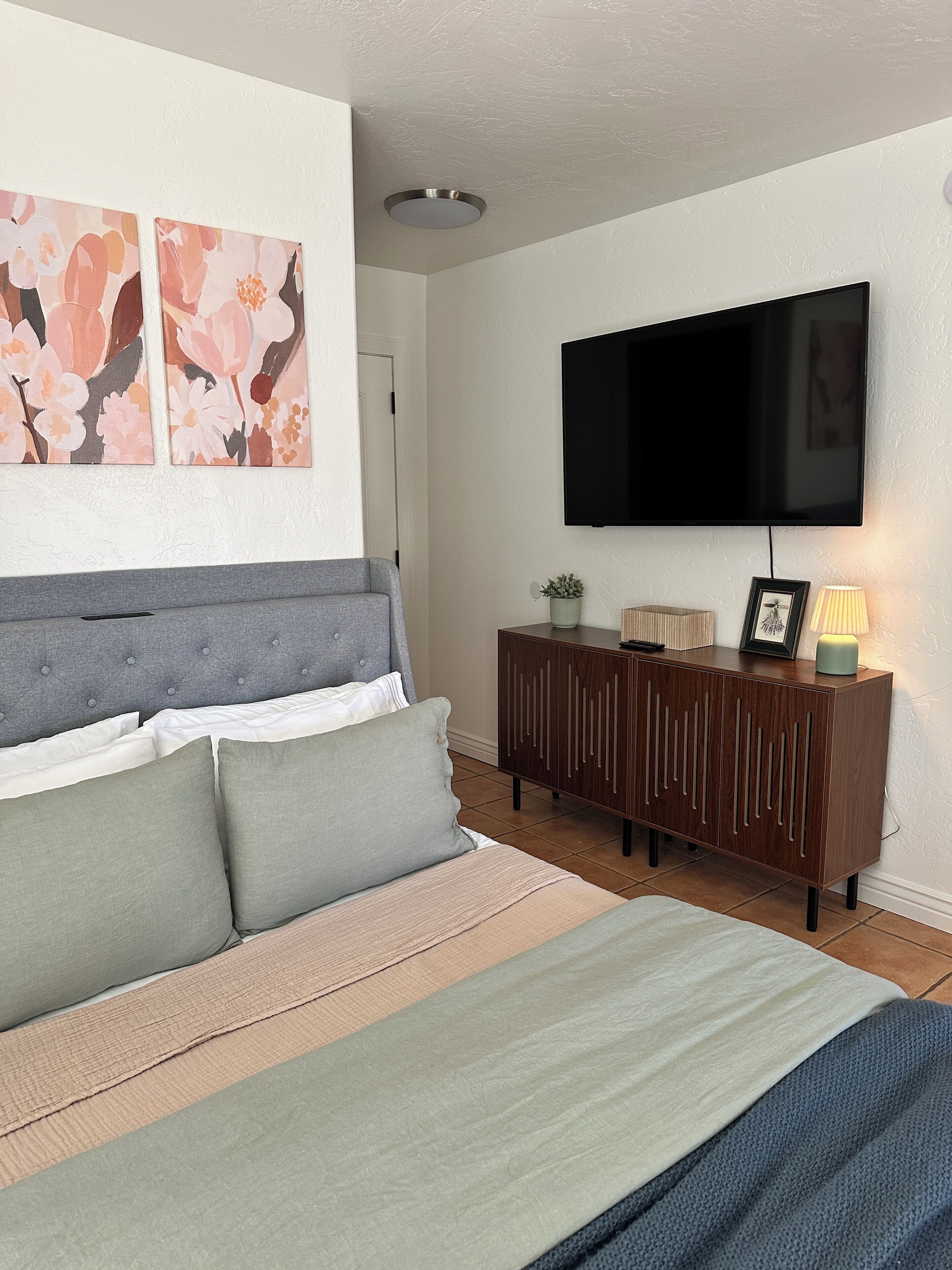 A bedroom with a bed, a TV mounted on the wall, and a cabinet with a lamp.