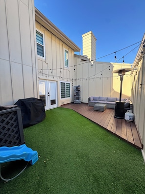 A backyard with artificial grass, a wooden deck, outdoor furniture, and string lights. - Spacious & Clean Home for 6 – Small Patio & Garage (Houston)
