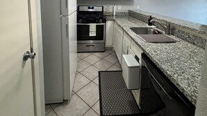 Une cuisine compacte avec des meubles blancs, un réfrigérateur en acier inoxydable, un four à micro-ondes, un four traditionnel, un lave-vaisselle et un plan de travail en granit.
