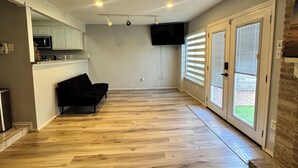 Un vaste espace de vie doté d’un plancher en bois, d’un canapé noir, d’un coin cuisine avec des meubles blancs, d’un téléviseur à écran plat fixé au mur et d’une porte-fenêtre coulissante donnant sur un patio.