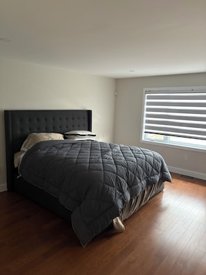 Un dormitorio con una cama de color oscuro, una ventana con persianas y suelo de madera.