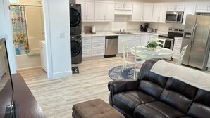 Une cuisine moderne avec des meubles blancs, un lave-vaisselle et un réfrigérateur. On y trouve un coin repas avec une table et des chaises en verre, ainsi qu’un canapé en cuir marron avec un pouf.
