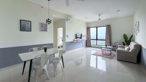 A modern living room with a dining area, a sofa, a TV, and a large window offering a city view. - 2BR Family Apartment in Melaka City (Malacca City)