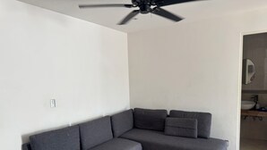 Ein modernes Wohnzimmer mit einer grauen Eckcouch, einem Deckenventilator und einem sichtbaren Badezimmer mit Spiegel und Waschbecken.