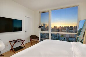 Una habitació d'hotel moderna amb un llit gran, una televisió penjada a la paret, una cadira i una vista a la ciutat a través de grans finestres.